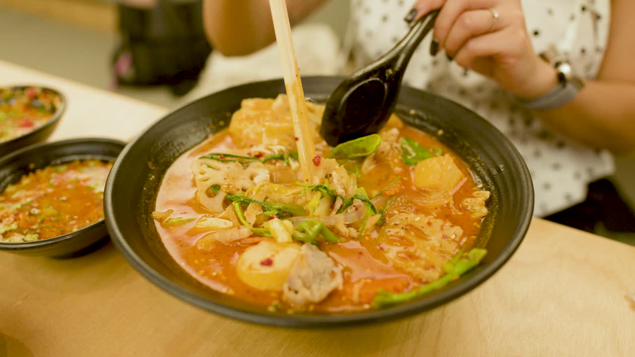 Close-up of woman using chopsticks and spoon to lift noodles from hot spicy Asian soup