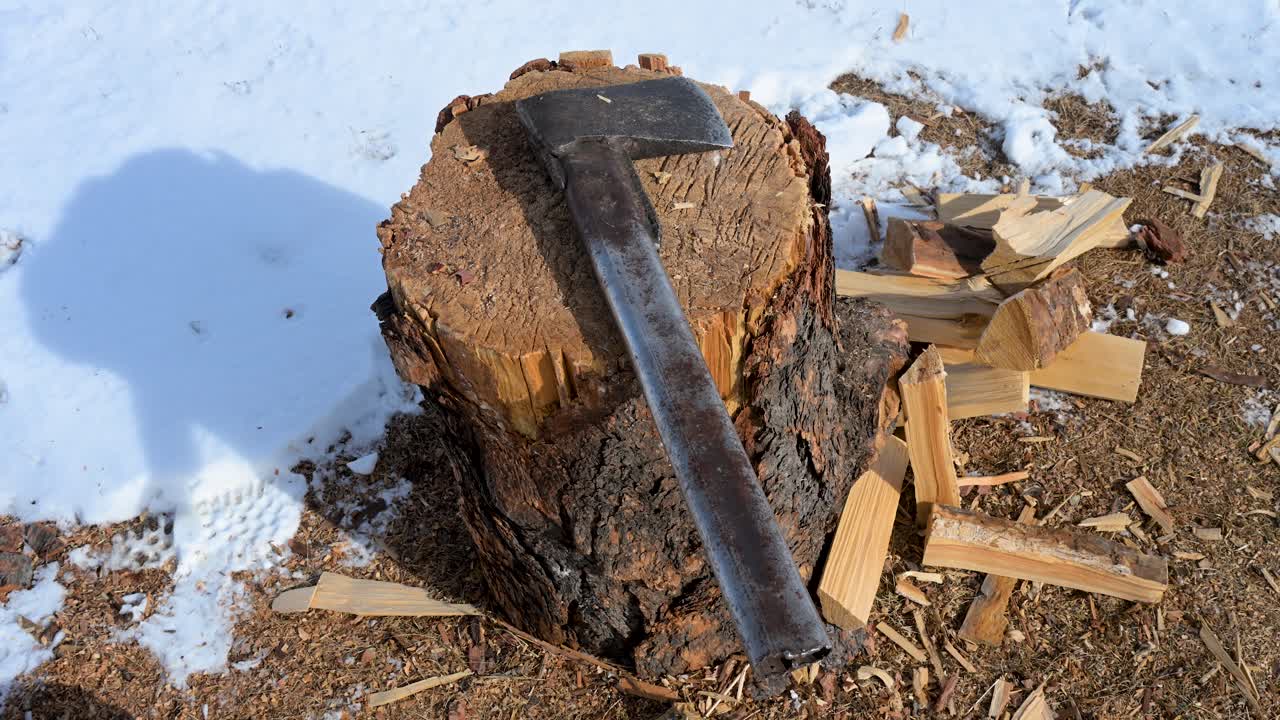 An axe rests on a chopping block, surrounded by freshly chopped kindling and wood chips in the snow. A scene of preparing firewood for heat and survival during a harsh winter