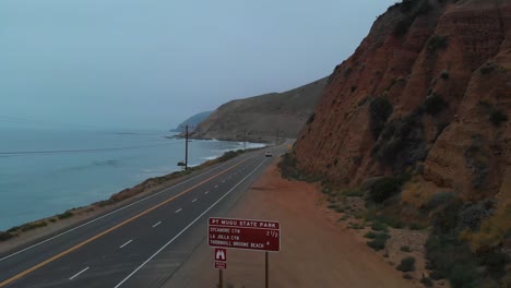 Rising-upward-drone-shot-as-car-pulls-out-onto-Pacific-Highway-One,-near-Malibu-on-a-grey-and-foggy-morning