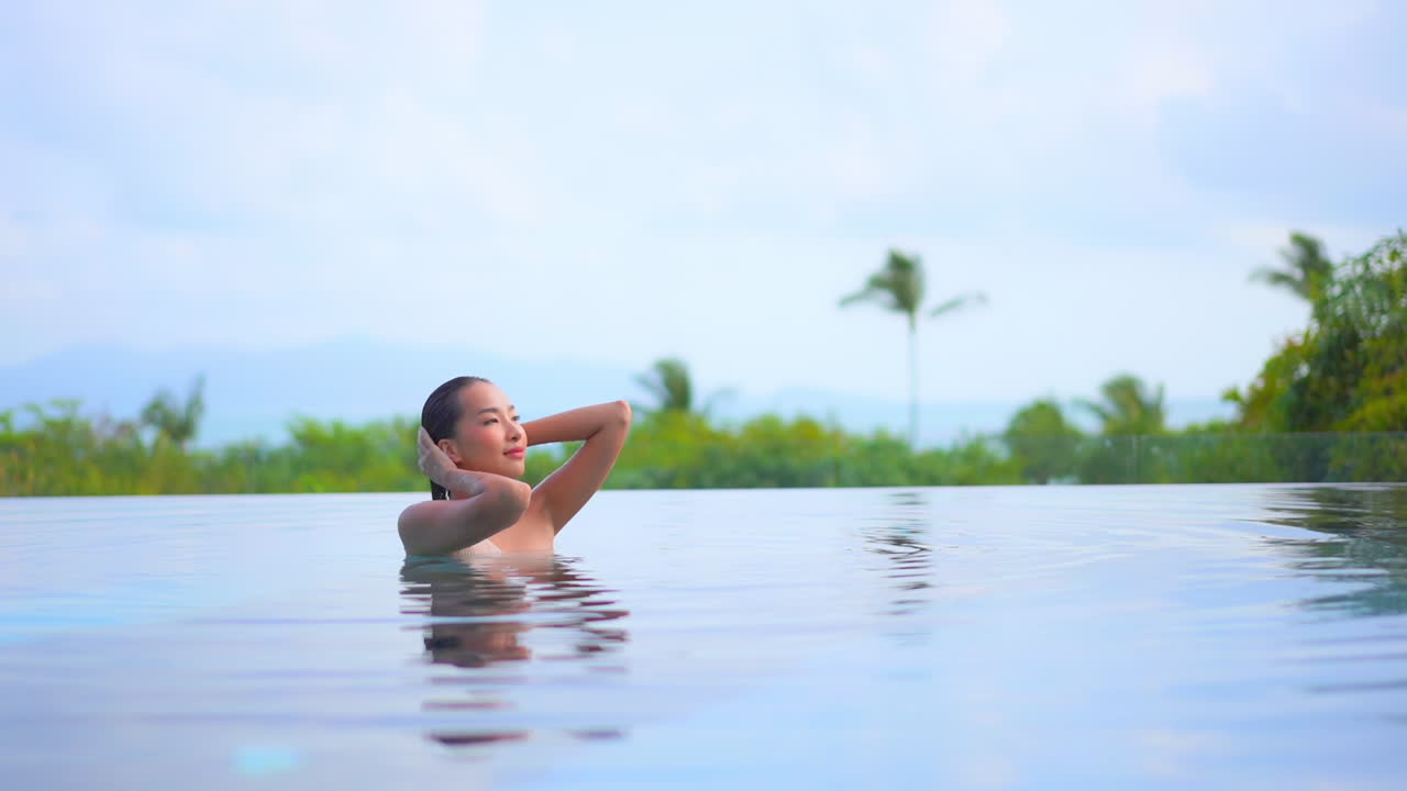 Junge, hübsche, exotische Frau in einem Infinity-Pool, die ihr nasses Haar richtet, mit tropischer Vegetation im Hintergrund, Zeitlupenaufnahme in voller Bildgröße.