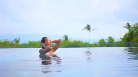 Young-pretty-exotic-woman-in-infinity-swimming-pool-fixing-her-wet-hair-tropical-vegetation-in-background,-full-frame-slow-motion