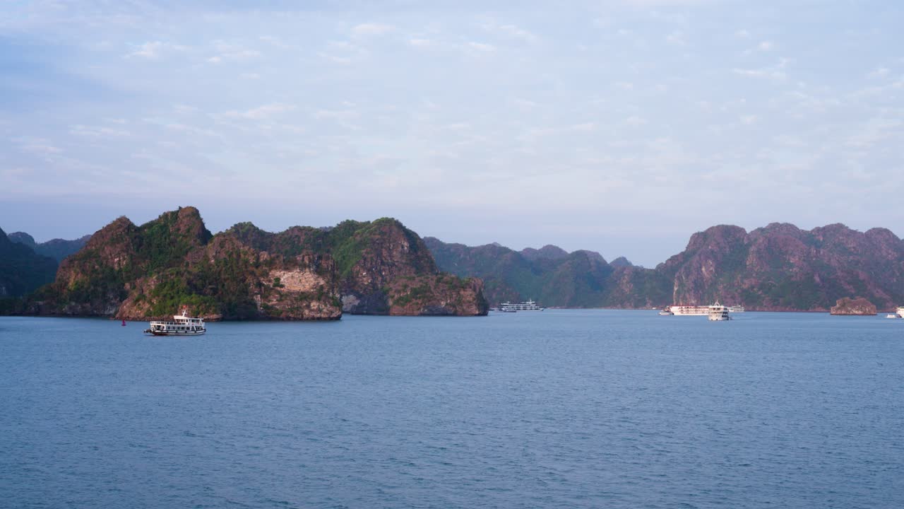 Limestone karst formations rise dramatically from the tranquil waters of Ha Long Bay, while traditional wooden cruise boats navigate between the towering islands under a soft, hazy blue sky