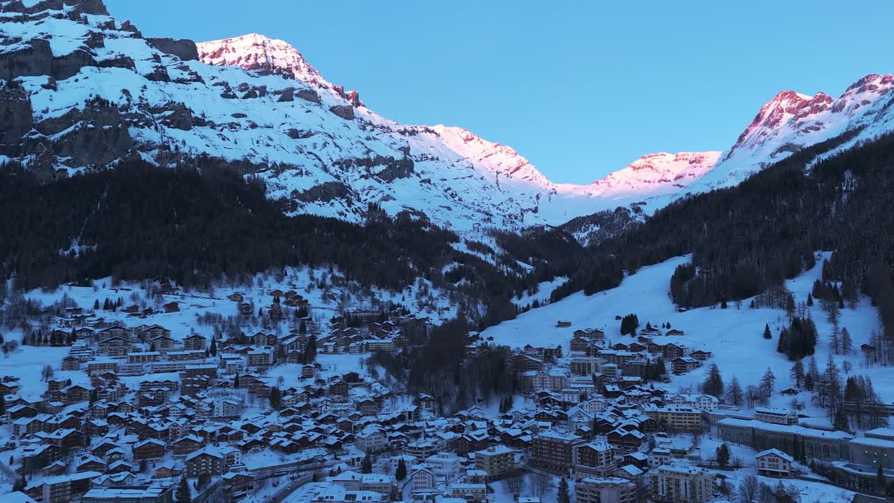 루이커바드의 겨울 풍경에서 드론 비행: 모든 것이 눈으로 여 있습니다. 저녁 태양은 산 꼭대기를 비추고 있습니다. 많은 휴양 주택과 호텔이있는 작은 산 마을을 볼 수 있습니다.