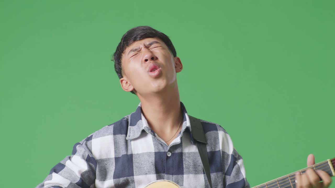 녹색 스크린 배경에서 기타를 연주하고 노래하는 젊은 아시아 십대 소년의 클로즈업
