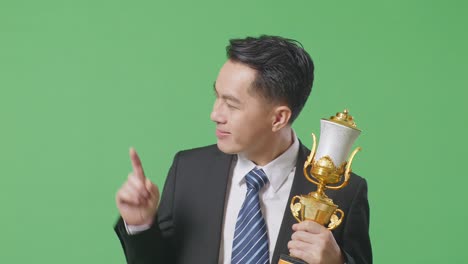 close up of asian business man in a suit and tie with a gold medal holding a gold trophy in his hands, smiling, and pointing to side on green screen background in the studio