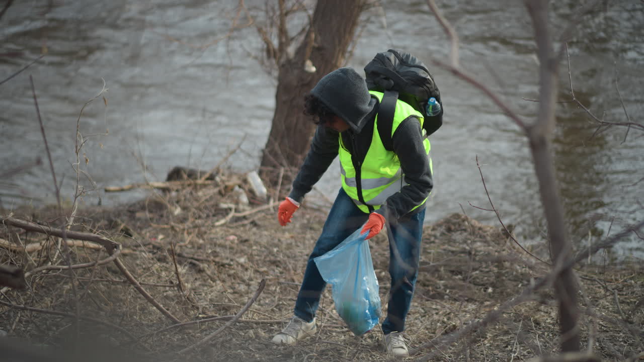 Volunteer in reflective vest and hoodie with backpack and gloves picks up litter along riverbank, placing trash into blue plastic bag during environmental cleanup to protect nature