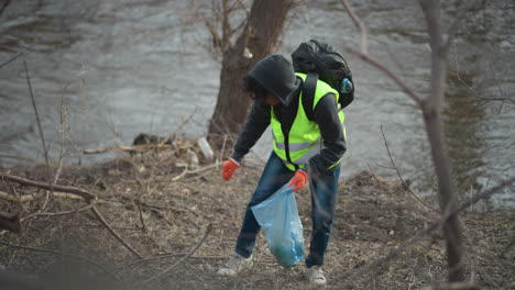 volunteer in reflective vest and hoodie with backpack and gloves picks up litter along riverbank, placing trash into blue plastic bag during environmental cleanup to protect nature