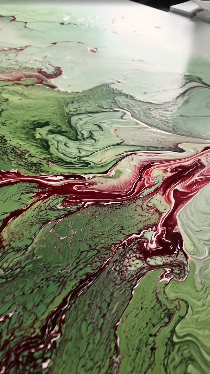 Rivoli rosso intenso si intrecciano attraverso un liquido verde pallido e bianco crema, mentre i riflessi lucidi rivelano una texture bagnata, bordi increspati e linee intricate che si muovono attraverso una composizione verticale ravvicinata