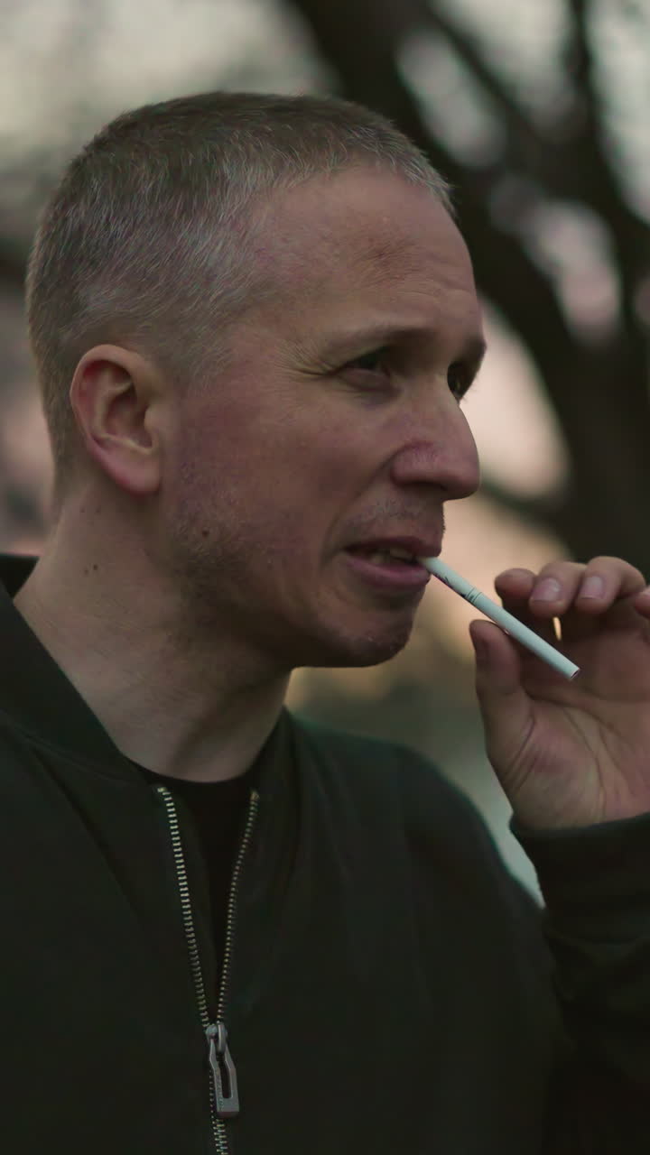 un hombre con una chaqueta verde está al aire libre al anochecer, sosteniendo un cigarrillo a su boca, profundamente en el pensamiento, con una vista borrosa de los coches que se mueven por detrás