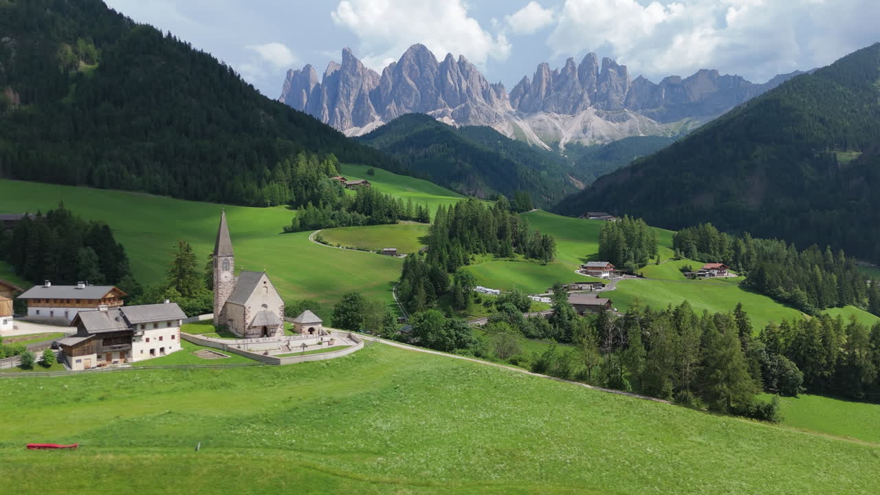 Scenic aerial drone shot of a church with the Dolomites mountains in the background, surrounded by green alpine meadows and traditional village houses