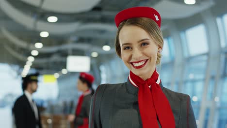 Retrato-De-La-Encantadora-Joven-Azafata-Rubia-En-Uniforme-Mirando-Y-Sonriendo-A-La-Cámara-Alegremente-En-La-Terminal-Del-Aeropuerto