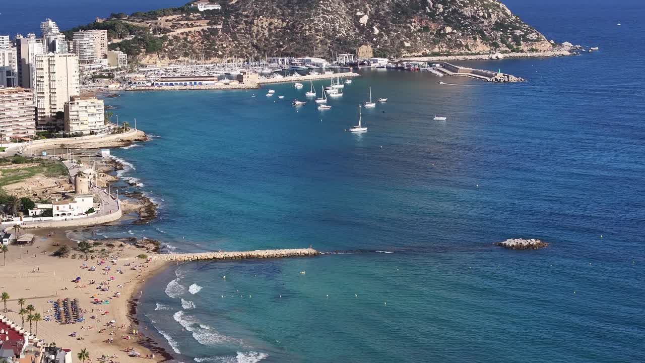 Calp beach with clear blue water, people lounging, and rocky breakwaters