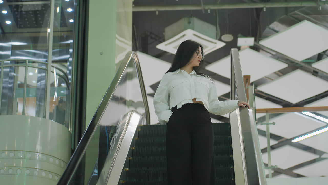 Professional woman wearing white shirt and black pants stands with hand in pocket while descending escalator in bright modern shopping mall, confidently gazing to the side with stylish posture
