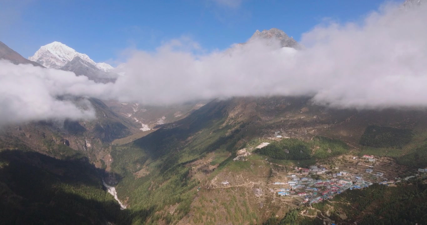 Ripresa lenta con drone di Namche Bazaar, Nepal, con uno strato di nuvole basse che attraversa l'inquadratura, separando la profonda valle dalle imponenti cime illuminate dal sole di Thamserku e Ama Dablam.