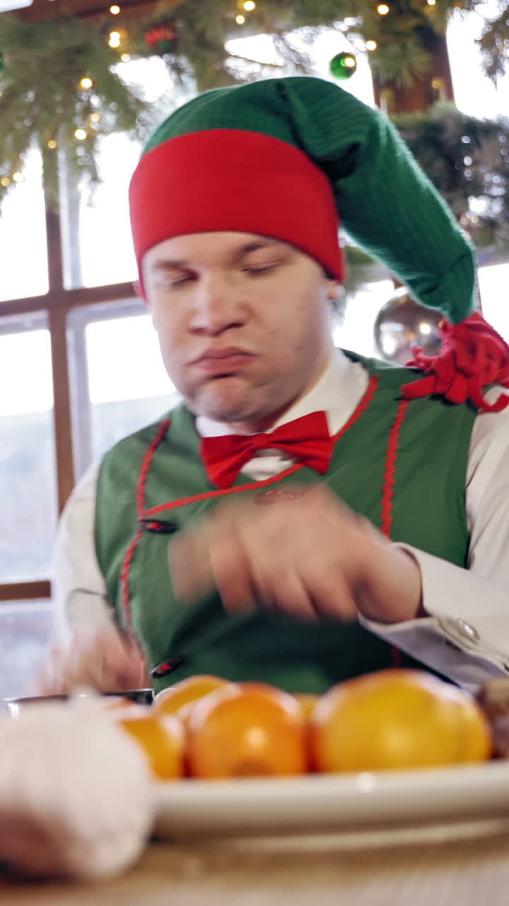 Hungry elf drinking at the table at Christmas. Male elf in green costume with hood eating at festive table on decorated background. Vertical video
