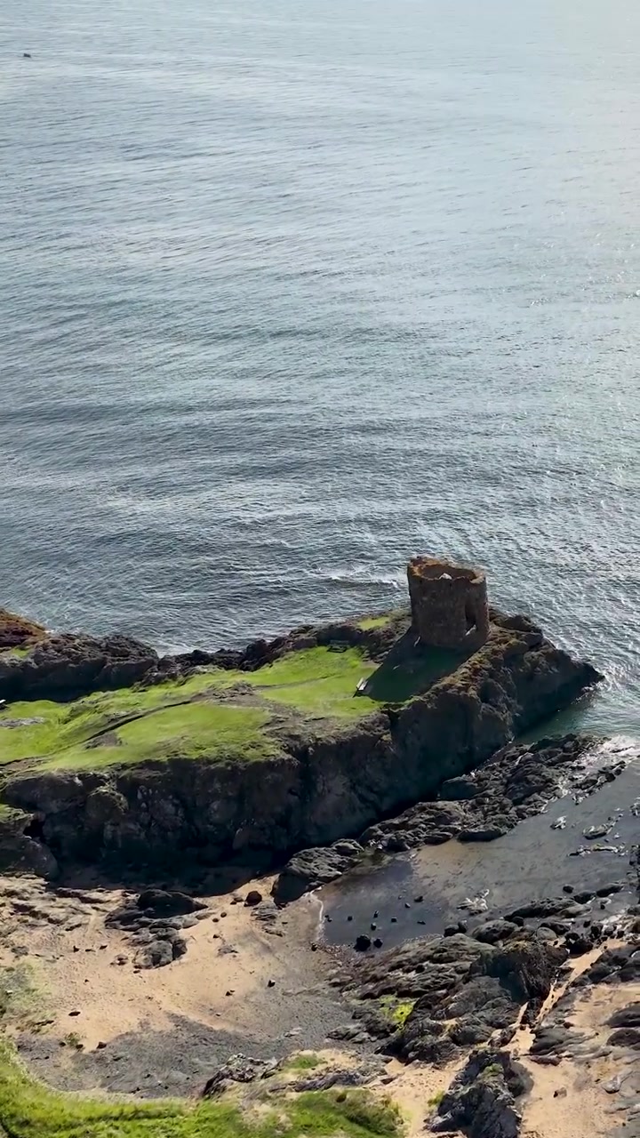 Un drone si innalza sopra una costa rocciosa, rovine antiche, tende e l'oceano sotto la luce naturale intensa del giorno.