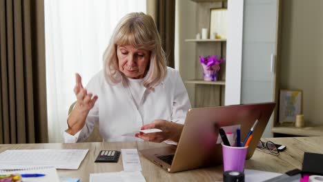 anciana ansiosa calculando impuestos en casa. mujer anciana preocupada tocando la cabeza y calculando impuestos mientras está sentada en la mesa con una computadora portátil en casa