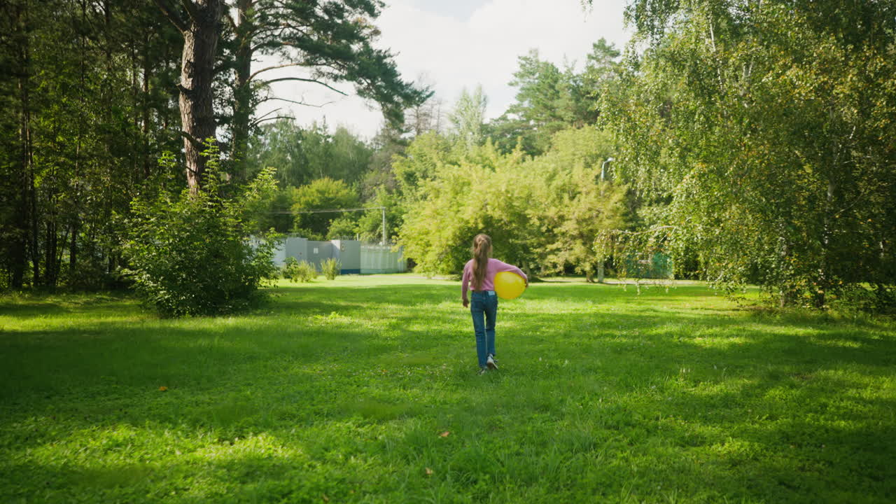 Back view of child walking through grassy field holding yellow balloon under right arm, surrounded by tall trees and soft sunlight filtering through leaves