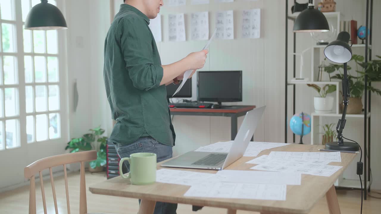 Creative Asian Designer Works On A Storyboard, Looks At His Sketches And Concepts, Choosing The Best Drawings For His Project. Video Editing, Comics Compilation, Application Ui Or Game Plot