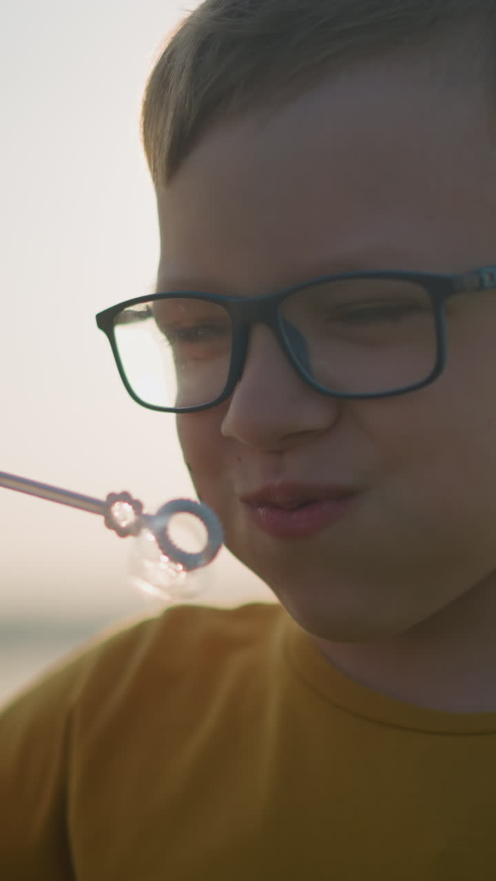 primer plano de un niño con una camisa amarilla y gafas mientras sumerge una varita en una botella de espuma y sopla burbujas en el aire al atardecer, capturando la alegría e inocencia de la infancia