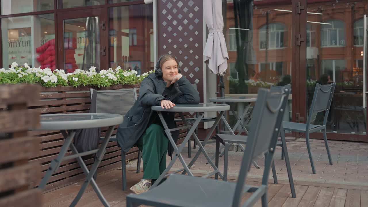 Donna caucasica seduta all'aperto nella terrazza di un bar, luce nuvolosa, fioriere in legno con fiori, sedie di metallo vuote, abito verde casual e cappotto scuro, sorriso rilassato e sguardo pensieroso, pavimentazione in pietra