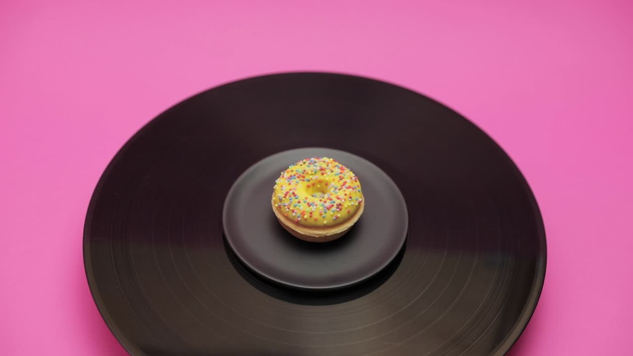 An American donut topped with yellow chocolate with  colorful crumbs turns in a circle on a black turntable. The shot from above