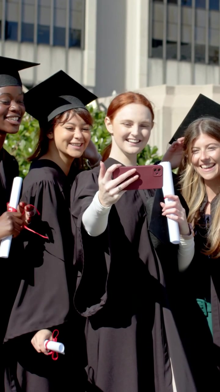 Vertical video: Raising pink phone, redhead grad taking selfie at campus with caps,gowns