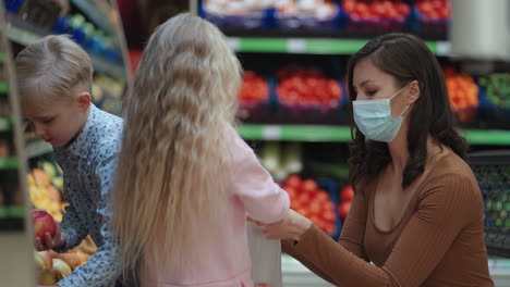 a mother in a protective mask with two children is buying groceries at the supermarket. buying food vegetables and fruits with children