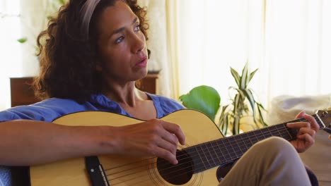 Frau-Mit-Gemischter-Abstammung-Spielt-Gitarre-Und-Singt,-Während-Sie-Zu-Hause-Auf-Der-Couch-Sitzt