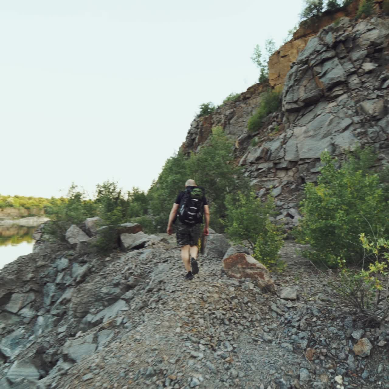 tourist with a backpack on his back is going along the bank of the lake on the background of high rocks and wild nature on a summer day