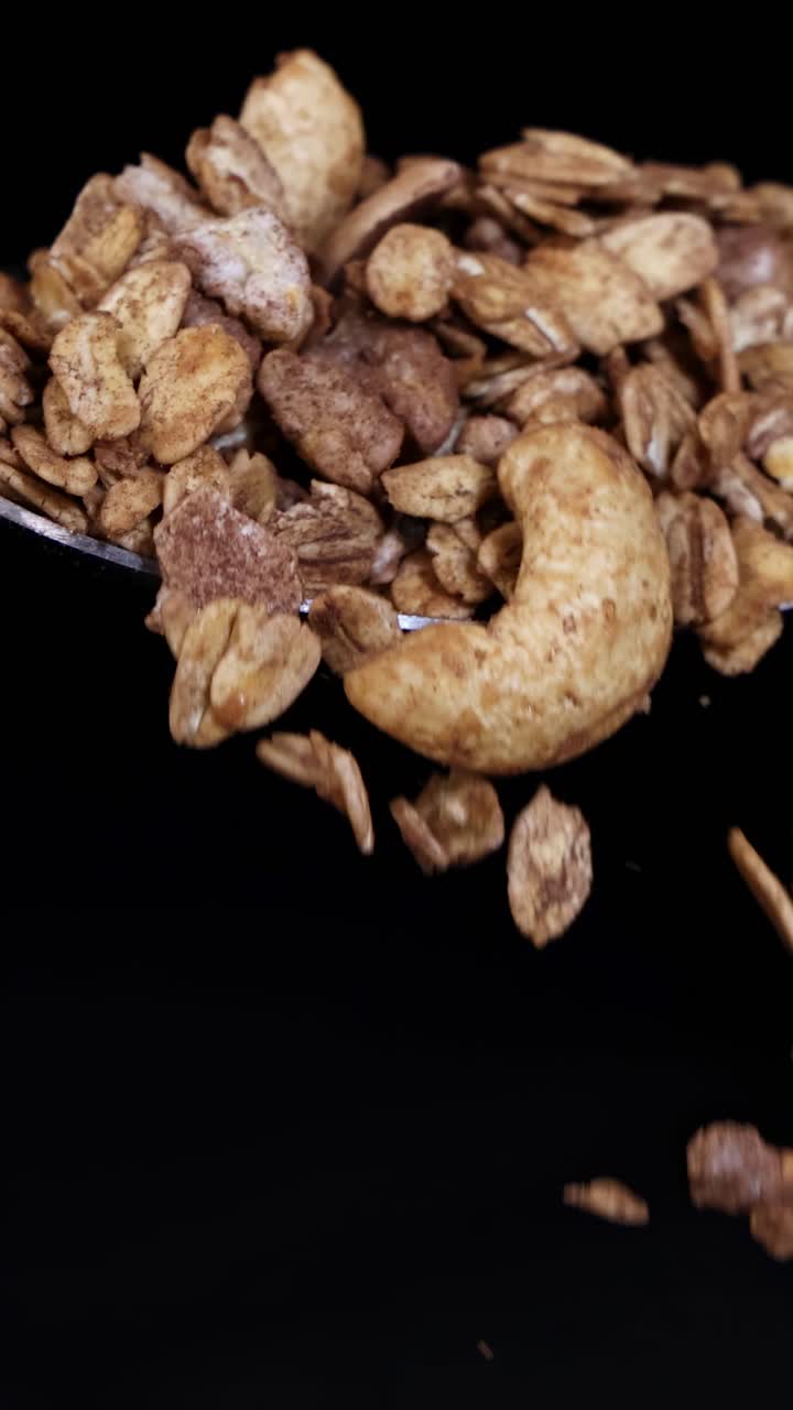Chicchi di granola scendono a cascata da un cucchiaio al rallentatore, su uno sfondo scuro, evidenziando la consistenza e il movimento.