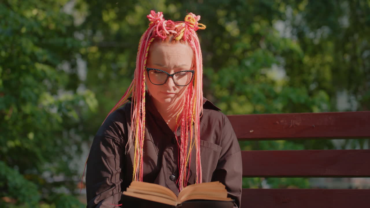 lector disfrutando de la luz del sol, persona con cabello vibrante absorta en libros en el exterior, mujer caucásica con trenzas de color rosa brillante concentrada intensamente en su libro entre la vegetación en una tarde soleada