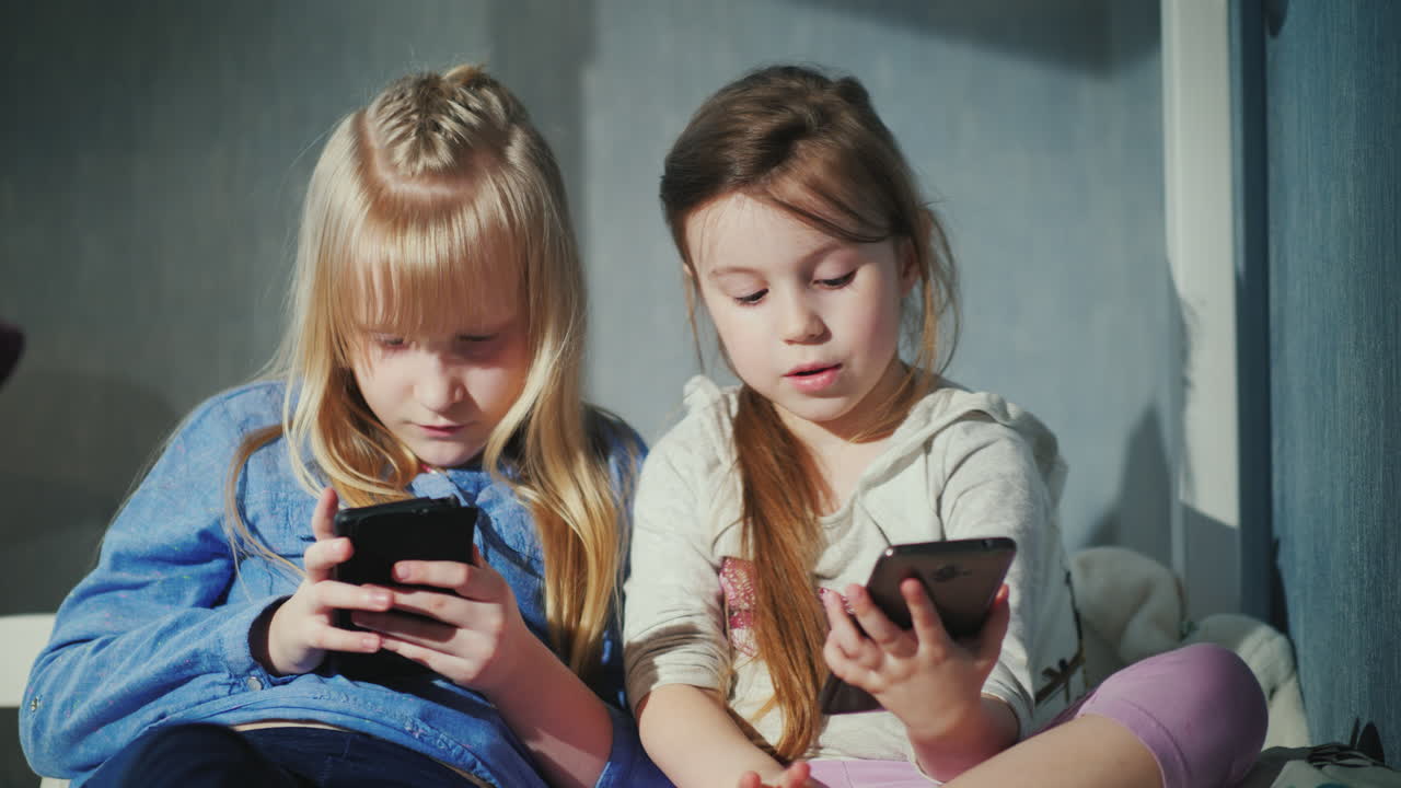 스마트폰으로 노는 두 소녀는 침실 4k 비디오에서 침대에 나란히 앉아 있습니다.