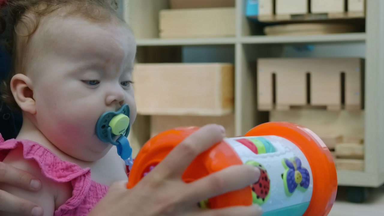 A baby girl chews on a colorful toy roller while held by an adult