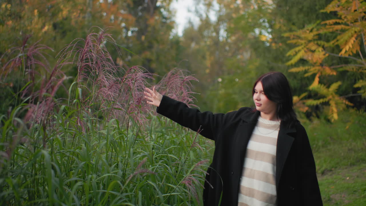 Young woman touches pine grove while walking through green park, extended arm brushing soft fronds, striped sweater under black coat, calm expression, warm autumn light over leaves