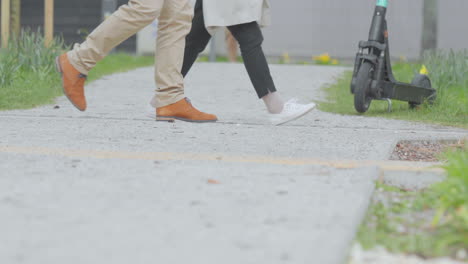 lower body view of two people walking on a concrete pathway, one wearing tan pants and orange shoes, the other in black pants and white shoes, a scooter parked to the side