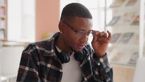 man with glasses works on laptop in library. young black guy with headphones adjusts spectacles sitting at computer in public hall. student technology