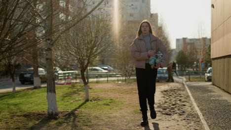 front view of a girl holding a skateboard while walking through an urban area at sunset. she is wearing a peach jacket, grey inner shirt, black pants, and black shoes