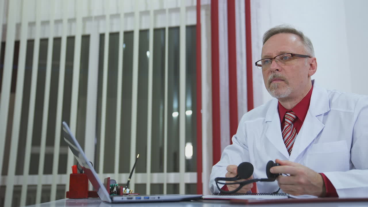 Doctor with headset working online. Professional senior physician talking to a patient through the laptop in his medical cabinet during quarantine.