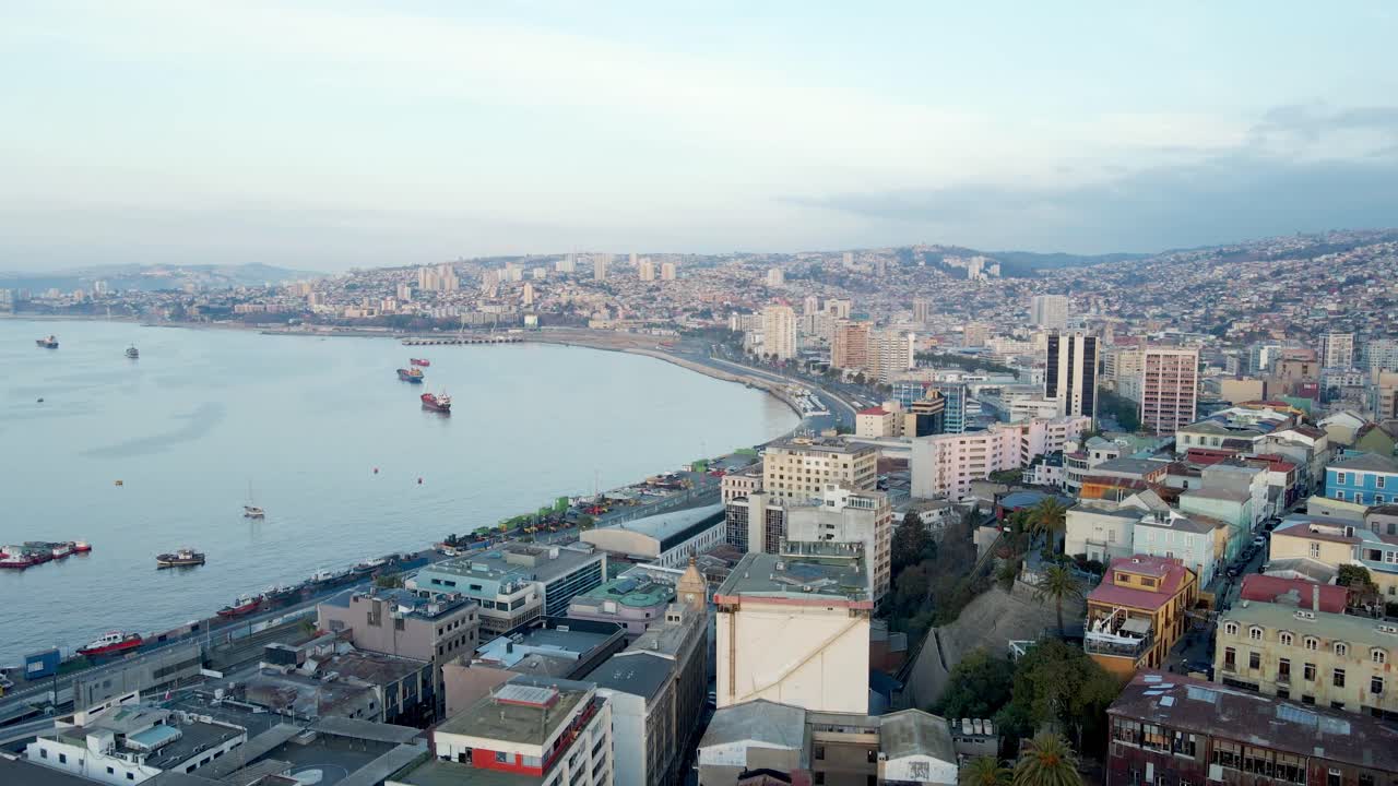 Ripresa aerea in avvicinamento degli edifici della città collinare di Valparaiso vicino al porto marittimo e navi che navigano vicino alla costa in una giornata nuvolosa, Cile