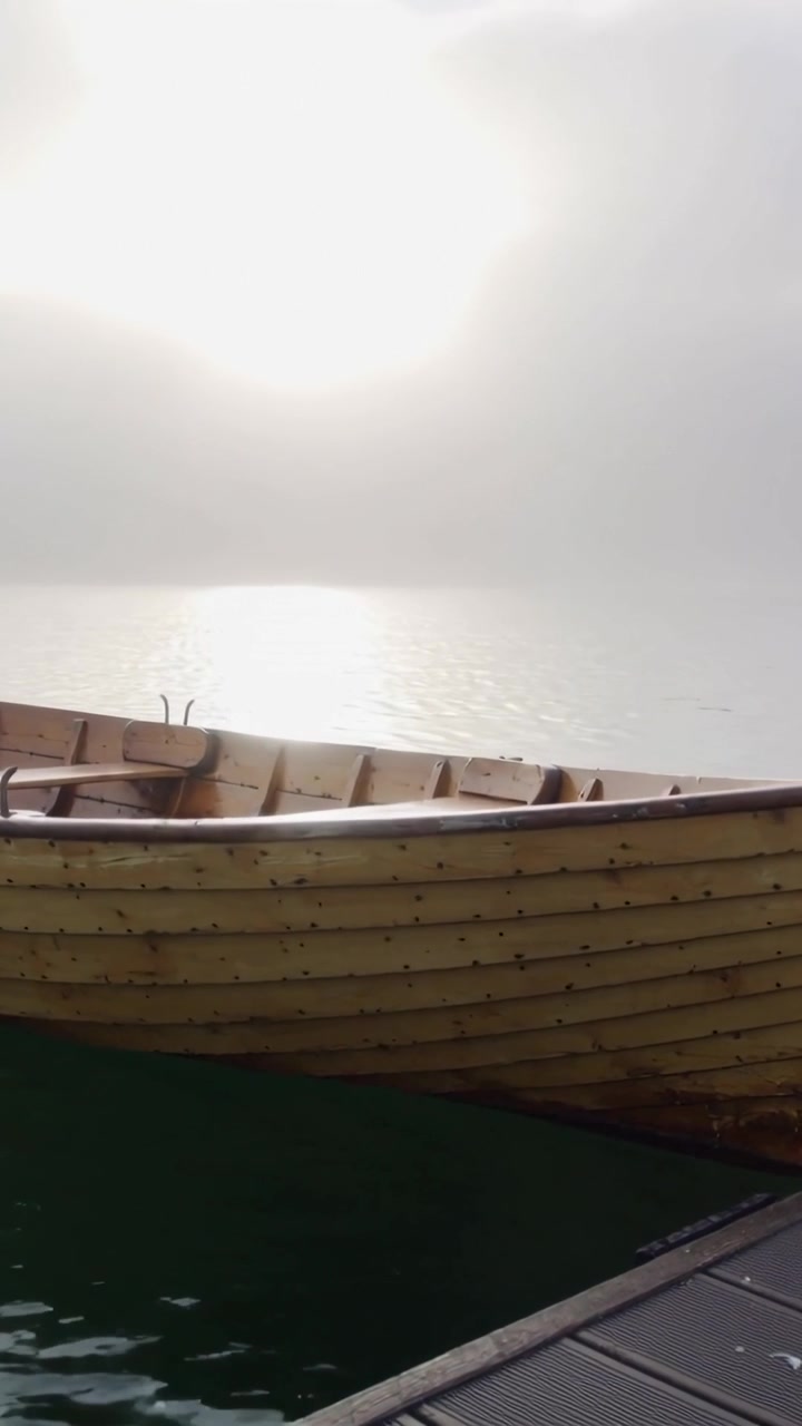 Vertikales Video: Sonne dringt durch Nebel, hölzernes Ruderboot schaukelt langsam am Steg, Wasser spiegelt sich