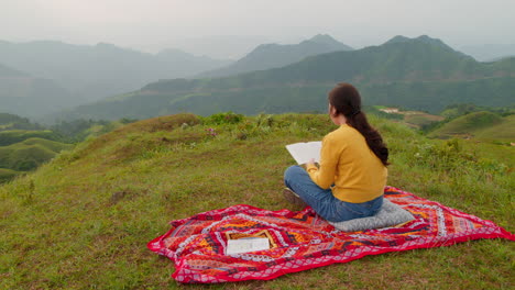 young woman reads while sitting on a red blanket at the top of a mountain