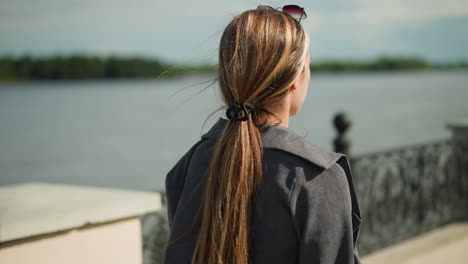 back view of a young lady walking near a bridge fence, with sunglasses resting on her head, the wind blows through her hair, with a blur view of river in the background