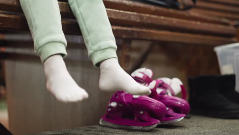 a close view of someone sitting on a bench, wearing white socks and green trousers, with pink ice skates and a black shoe by the side, the person playfully dangles their legs