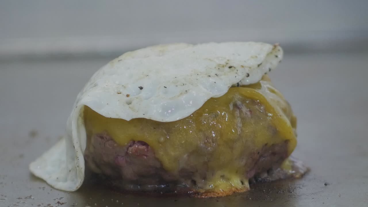 primer plano de una hamburguesa con queso en la parrilla de un restaurante mientras se coloca un huevo frito encima