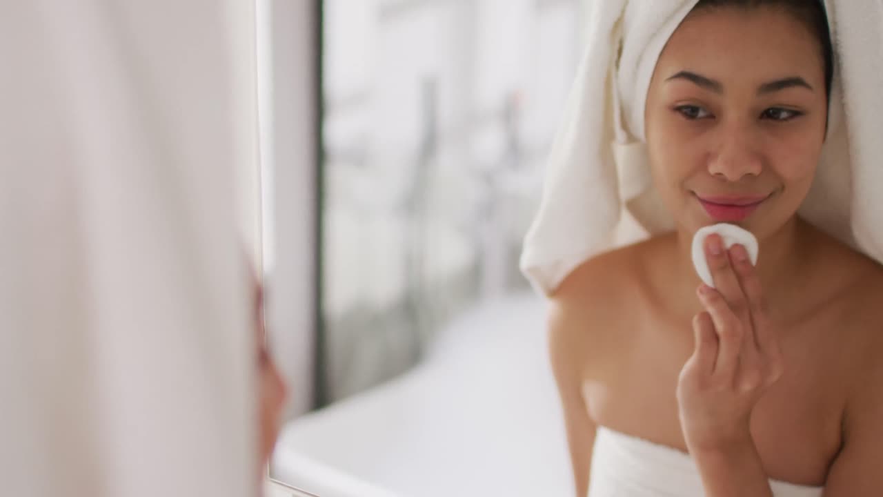 Video of portrait of smiling biracial woman with towel on hair cleansing face in bathroom