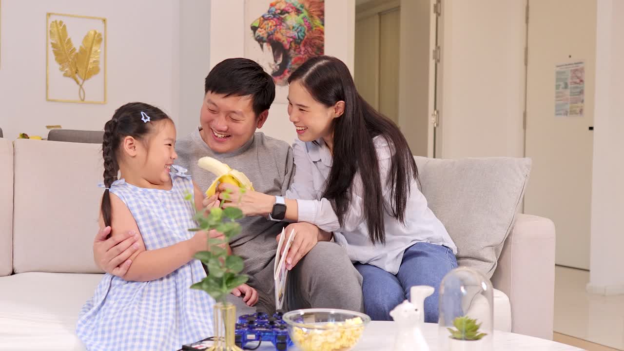 Smiling parents and daughter share laughter, play, and bananas on a bright living room sofa