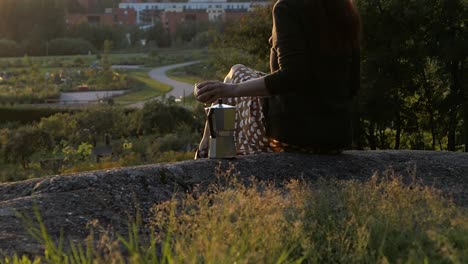 Woman-sitting-on-rock-at-sunset-golden-hour-backlight,-rising-crane-shot