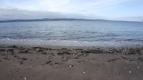 Calm-waves-on-a-beach-looking-across-the-water-to-an-island-|-Edinburgh,-Scotland-|-4k-at-30-fps