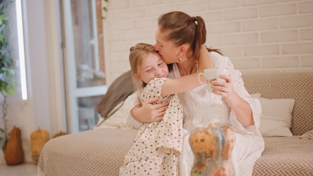 Weiße Mutter und Tochter umarmen sich mit Tasse, Kürbiskuchen daneben auf Holztisch, spielerische Frühstücksszene, sonnenbeschienene Ziegelwand und gemütliches Sofa, Mutter bietet warmes Getränk an, während Tochter kichert, idyllisch.